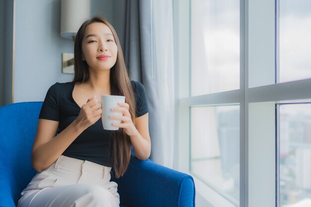 肖像画の美しい若いアジアの女性は、リビングルームのソファーの椅子にコーヒーカップを保持します。