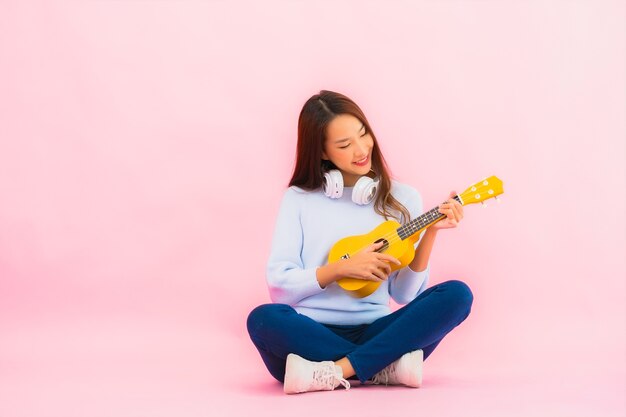 Portrait beautiful young asian woman play ukulele on pink color isolated wall