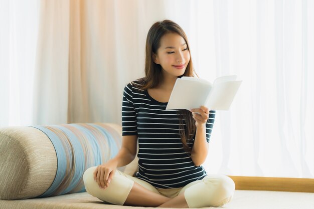 Книга чтения женщины портрета красивая молодая азиатская внутри на софе в зоне живущей комнаты