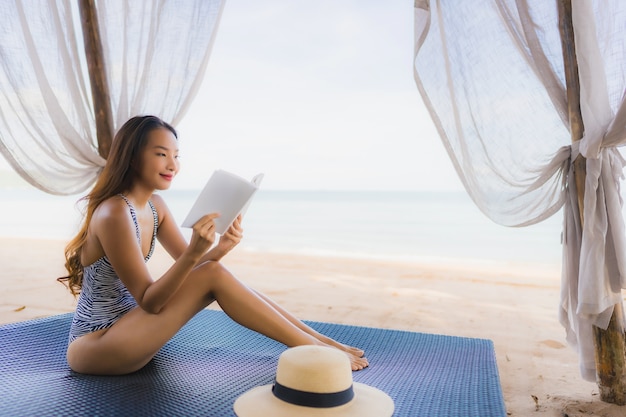행복 한 미소로 초상화 아름 다운 젊은 아시아 여자 읽기 책 레저 해변 바다 바다 라운지 침대의 자에서 휴식