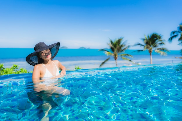 La bella giovane donna asiatica del ritratto si rilassa nella piscina all'aperto di lusso nell'oceano quasi del mare della spiaggia della località di soggiorno dell'hotel