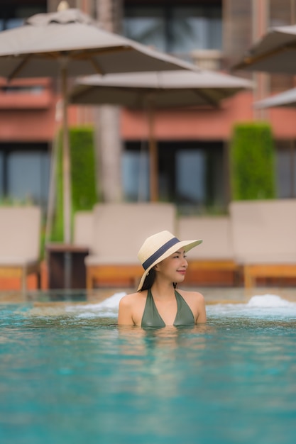 La bella giovane donna asiatica del ritratto si rilassa nella piscina all'aperto di lusso nell'oceano quasi del mare della spiaggia della località di soggiorno dell'hotel