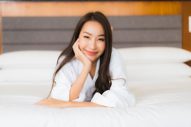 肖像画の美しい若いアジア女性は寝室のインテリアでベッドの上の笑顔をリラックスします。