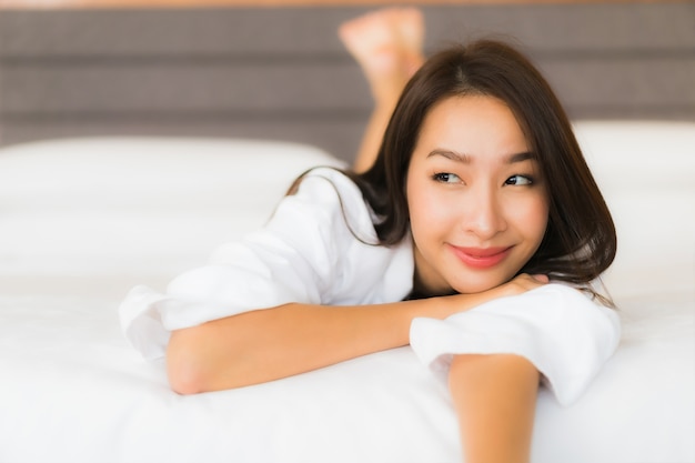 肖像画の美しい若いアジア女性は寝室のインテリアでベッドの上の笑顔をリラックスします。