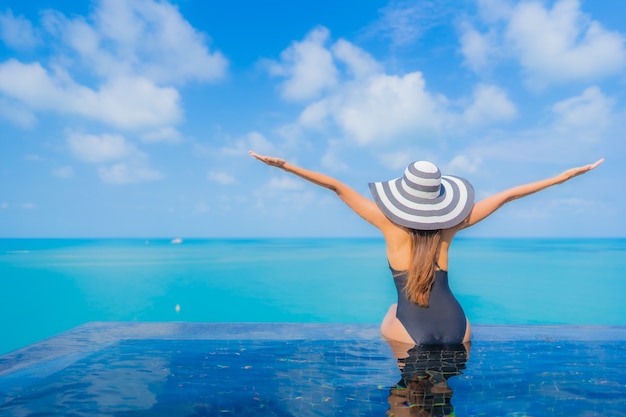 Foto gratuita la bella giovane donna asiatica del ritratto si rilassa il tempo libero di sorriso intorno alla piscina all'aperto nell'hotel della località di soggiorno con la vista dell'oceano del mare