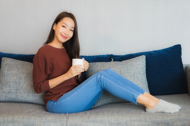 Женщина портрета красивая молодая азиатская ослабляет улыбку на софе в интерьере живущей комнаты