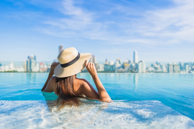 Bella giovane donna asiatica del ritratto che si rilassa intorno alla piscina all'aperto con la vista della città