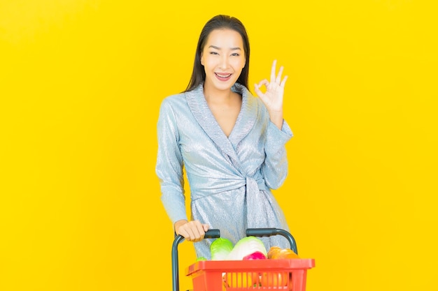 Улыбка женщины портрета красивая молодая азиатская с продуктовой корзиной от супермаркета на желтой стене