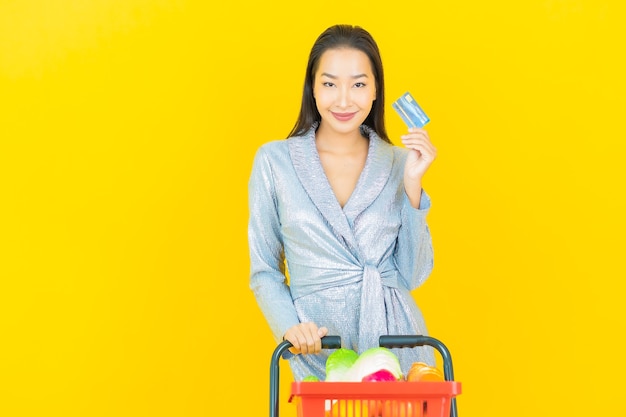 Free photo portrait beautiful young asian woman smile with grocery basket from supermarket on yellow wall