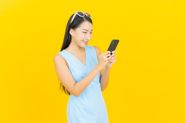 Улыбка женщины портрета красивая молодая азиатская с умным мобильным телефоном на стене желтого цвета