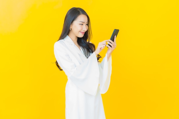Портрет красивой молодой азиатской улыбки женщины с умным мобильным телефоном на желтой стене
