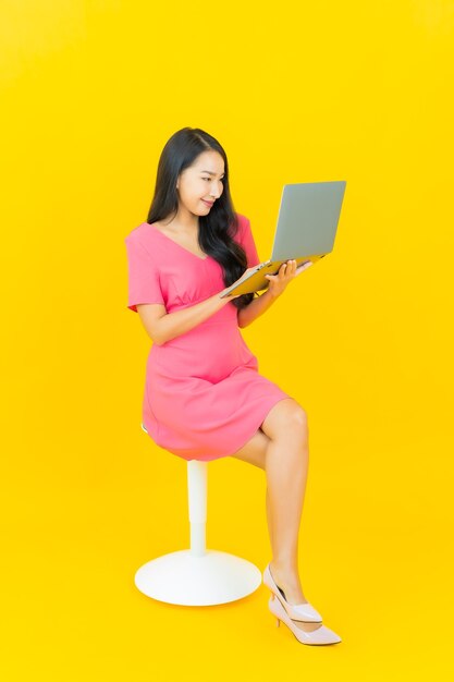 Portrait beautiful young asian woman smiles with computer laptop on yellow isolated wall