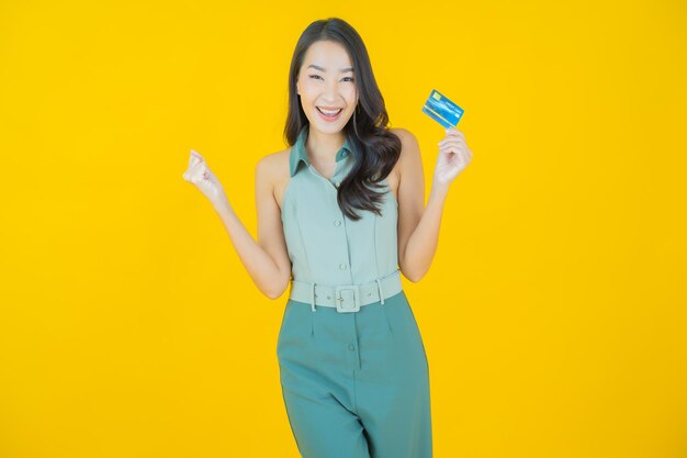 노란 벽에 신용카드를 들고 웃는 아름다운 젊은 아시아 여성의 초상화