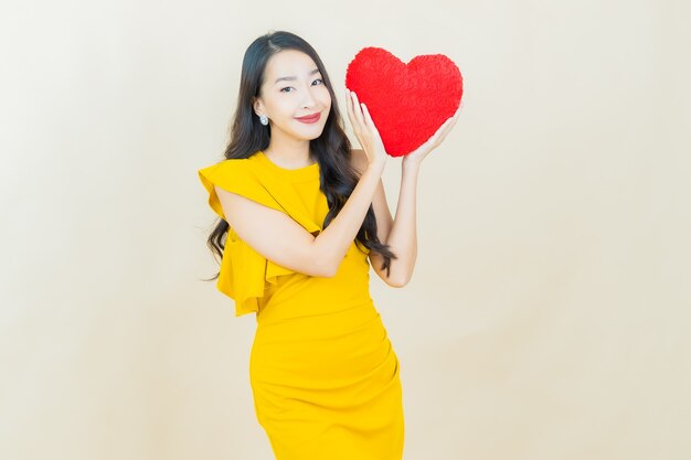 베이지색 벽에 심장 베개 모양으로 미소 짓는 아름다운 젊은 아시아 여성