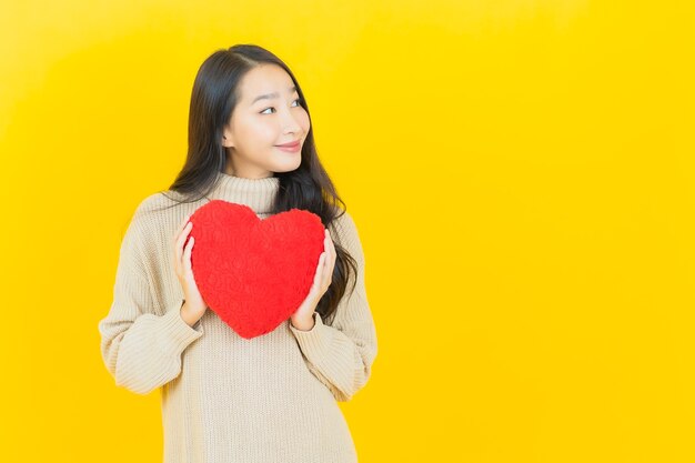 노란색 벽에 심장 베개 모양 세로 아름 다운 젊은 아시아 여자 미소