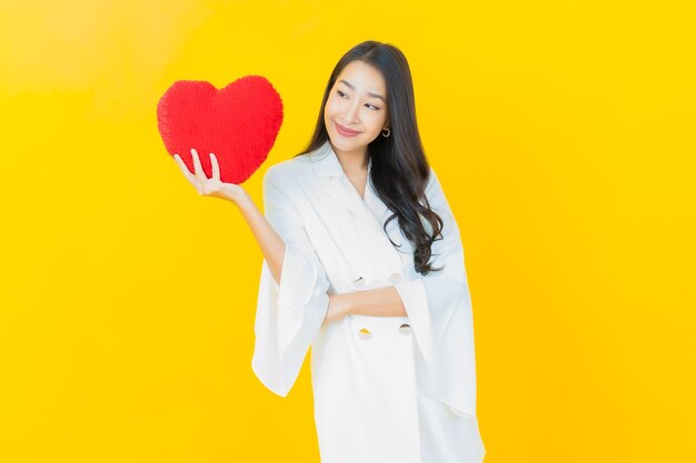 Free photo portrait of beautiful young asian woman smiles with heart pillow shape on yellow wall