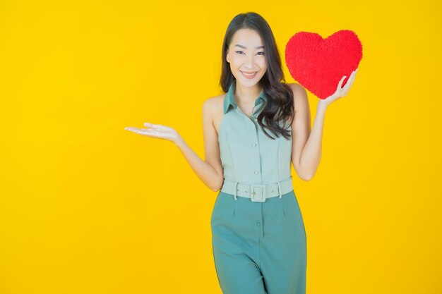 Portrait of beautiful young asian woman smiles with heart pillow shape on yellow wall