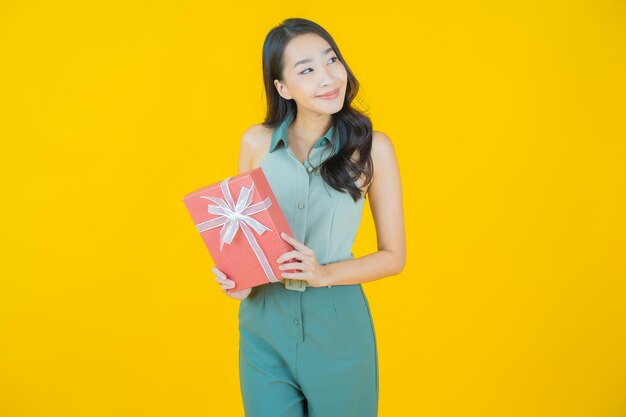 노란 벽에 빨간 선물 상자를 들고 웃는 아름다운 젊은 아시아 여성의 초상화