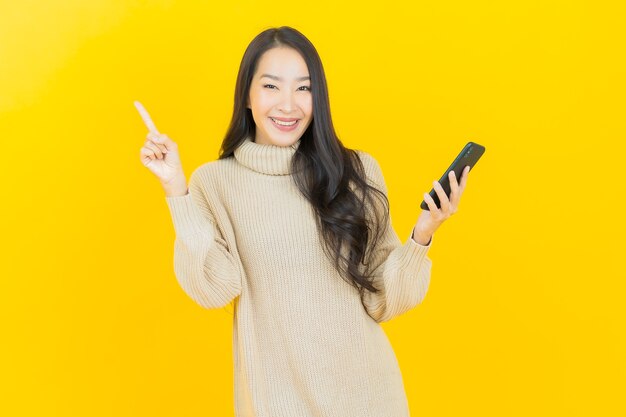 黄色の壁にスマートな携帯電話で笑顔美しい若いアジアの女性の肖像画