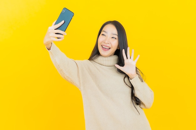 노란색 벽에 스마트 휴대 전화와 세로 아름 다운 젊은 아시아 여자 미소