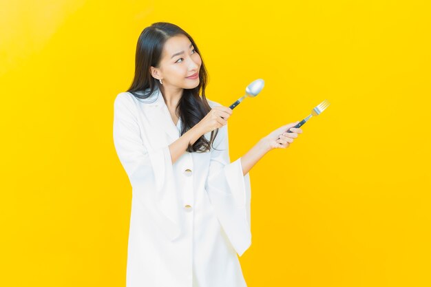 Portrait of beautiful young asian woman smiles with spoon and fork on yellow wall