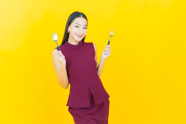 Free photo portrait beautiful young asian woman smiles with spoon and fork on yellow wall