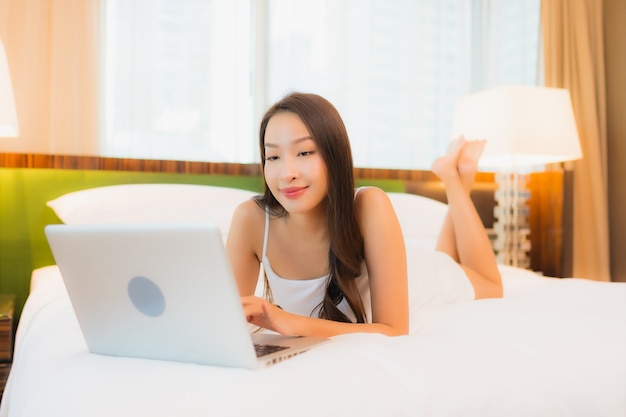 Компьтер-книжка компьютера пользы женщины портрета красивая молодая азиатская на кровати в интерьере спальни