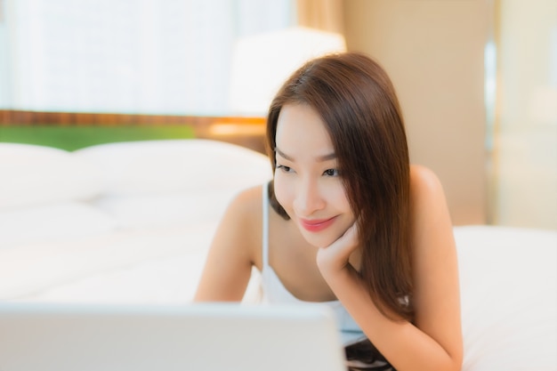 Компьтер-книжка компьютера пользы женщины портрета красивая молодая азиатская на кровати в интерьере спальни