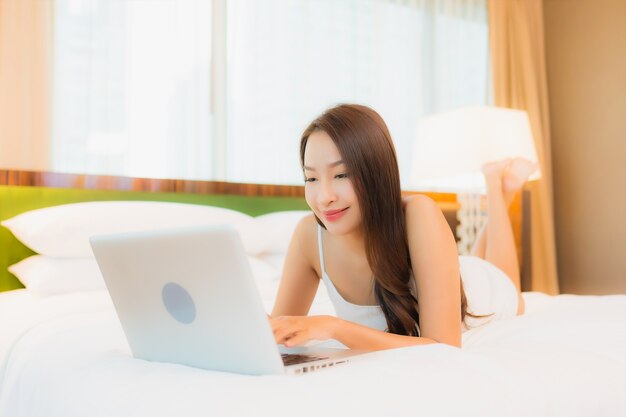 肖像画の美しい若いアジア女性は寝室のインテリアのベッドの上のコンピューターのラップトップを使用します。