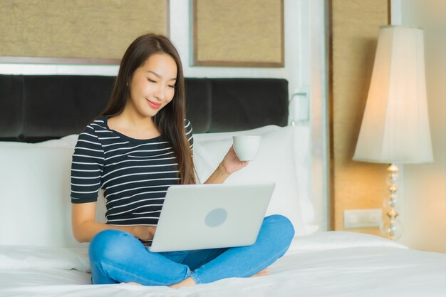 肖像画の美しい若いアジア女性は寝室のインテリアのベッドの上のコンピューターのラップトップを使用します。