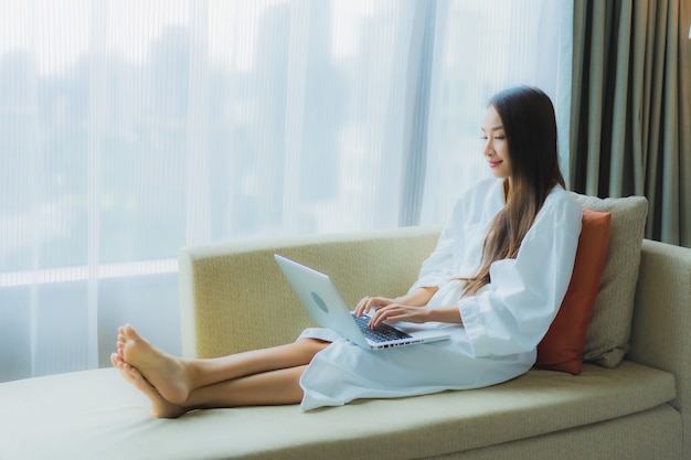 肖像画の美しい若いアジアの女性は、リビングルームエリアのソファーにコンピューターラップトップを使用します。