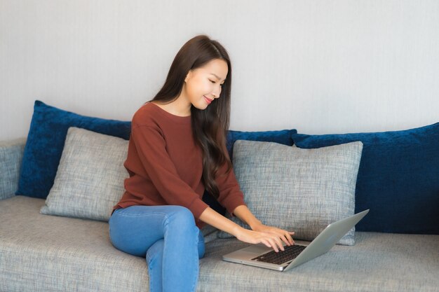 肖像画の美しい若いアジア女性は、リビングルームのインテリアのソファーにコンピューターラップトップを使用します。