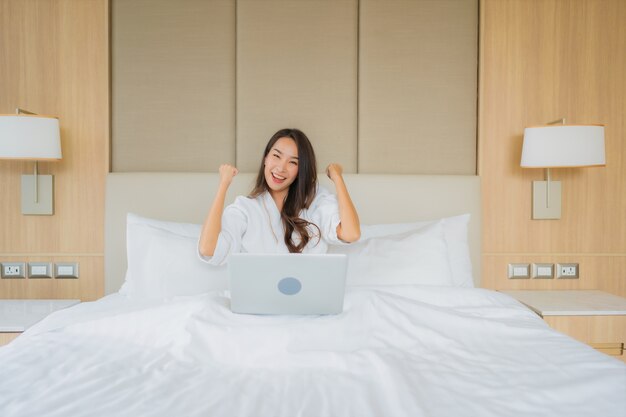 肖像画の美しい若いアジア女性は寝室でラップトップコンピューターを使用します。