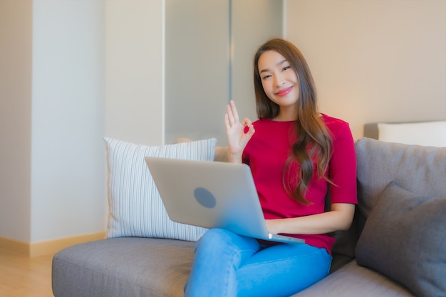 Портативный компьютер пользы женщины портрета красивый молодой азиатский на софе в жилой зоне