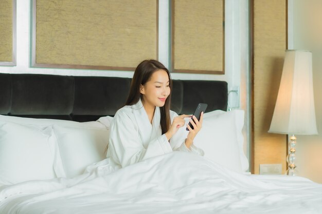 Мобильный телефон пользы женщины портрета красивой молодой азиатской умный на кровати в интерьере спальни