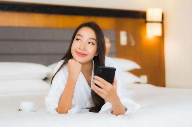 肖像画の美しい若いアジア女性は寝室のインテリアでベッドの上のスマートな携帯電話を使用します。