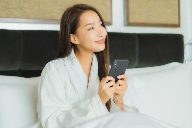 肖像画の美しい若いアジア女性は寝室のインテリアでベッドの上のスマートな携帯電話を使用します。