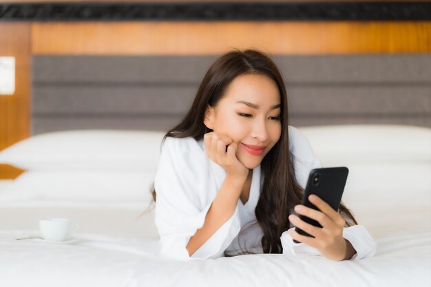 Мобильный телефон пользы женщины портрета красивой молодой азиатской умный на кровати в интерьере спальни