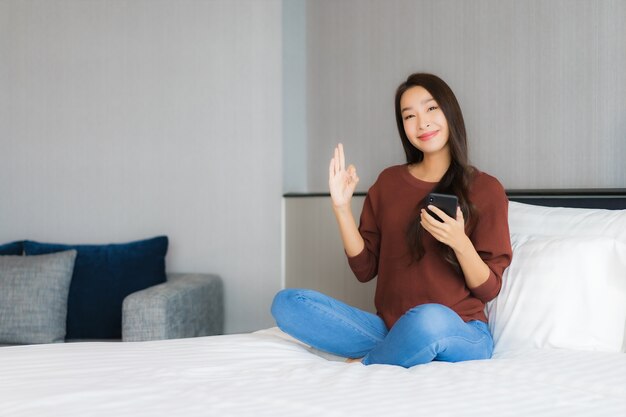 肖像画の美しい若いアジア女性は寝室のインテリアでベッドの上のスマートな携帯電話を使用します。