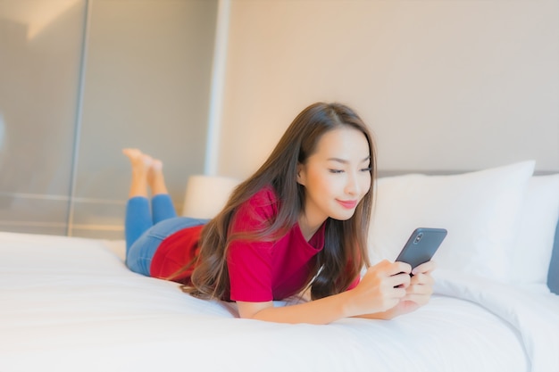 美しい若いアジア女性の肖像画はベッドの上のスマートな携帯電話を使用します。