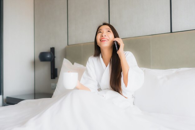 肖像画の美しい若いアジア女性は寝室でスマートな携帯電話を使用します。