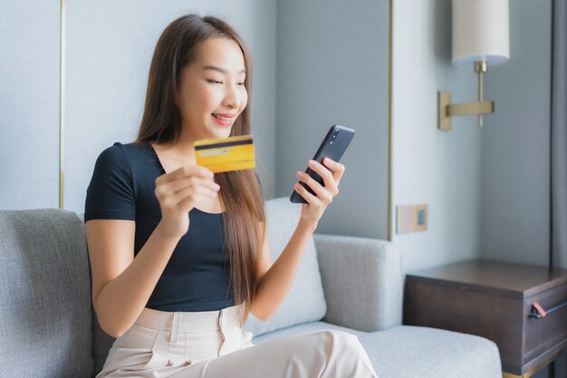 거실에서 소파에 신용 카드로 세로 아름 다운 젊은 아시아 여자 사용 스마트 휴대 전화 또는 노트북