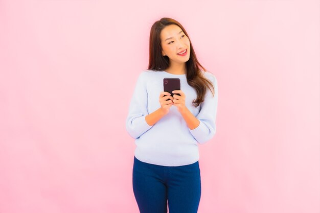 肖像画美しい若いアジアの女性はピンク色の隔離された壁にスマート携帯電話を使用します