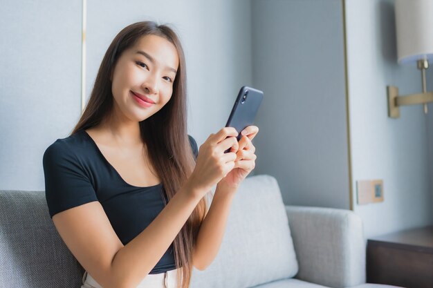 肖像画の美しい若いアジア女性はリビングルームエリアのソファーにスマートな携帯電話を使用します。
