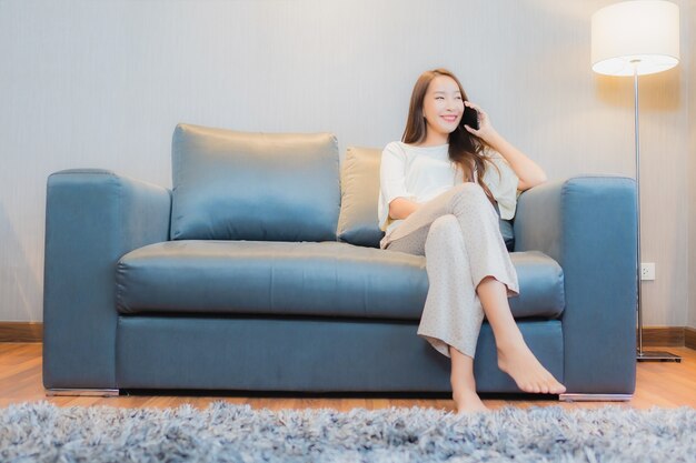 Мобильный телефон пользы женщины портрета красивой молодой азиатской умный на софе в интерьере живущей комнаты