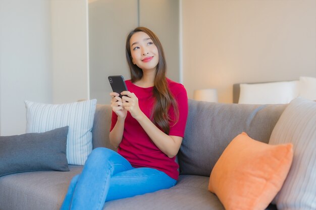肖像画の美しい若いアジア女性はソファーでスマートな携帯電話を使用します。