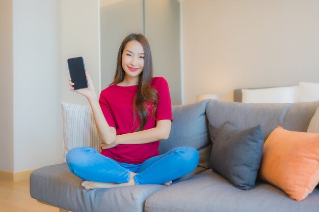 肖像画の美しい若いアジア女性はソファーでスマートな携帯電話を使用します。