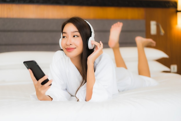 La bella giovane donna asiatica del ritratto utilizza il telefono cellulare astuto con la cuffia per ascoltare la musica sul letto nell'interno della camera da letto