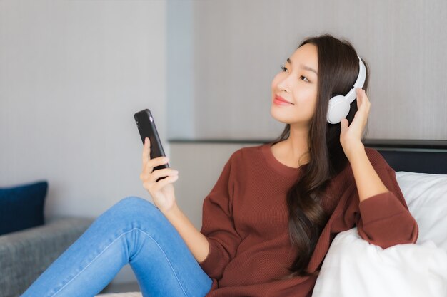 肖像画の美しい若いアジア女性は寝室のインテリアのベッドで音楽を聴くためのヘッドフォンでスマートな携帯電話を使用します。