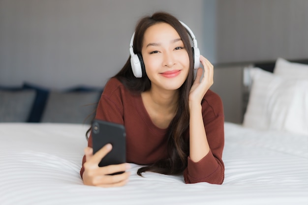 Portrait beautiful young asian woman use smart mobile phone with headphone for listen music on bed in bedroom interior